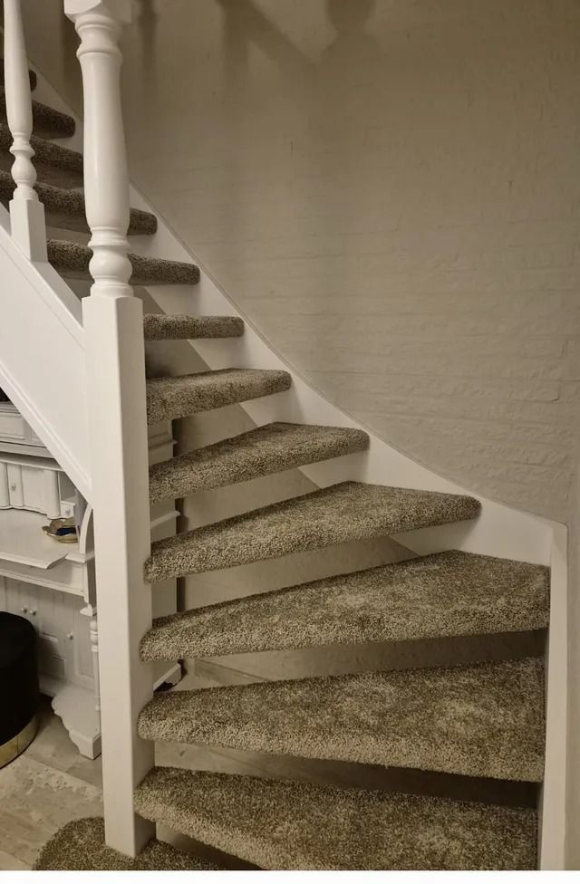 Een gebogen trap met tapijtbekleding, een witte leuning en een beige muur in een woninginterieur.