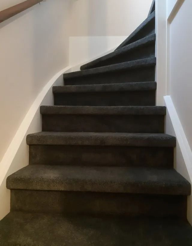 Een met tapijt beklede trap met witte muren, die in een smalle gang naar rechts omhoog buigt.
