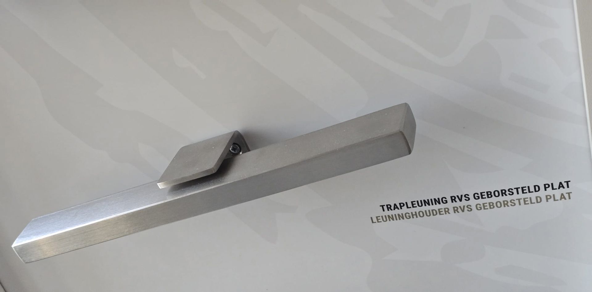 Een schuin geplaatst metalen bord aan de muur met kleine zwarte tekst en daarboven een grijze, scharnierachtige beugel.