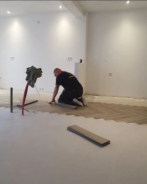 Een arbeider knielt op de vloer en legt houten vloerdelen in een lege witte ruimte, met gereedschap in de buurt.