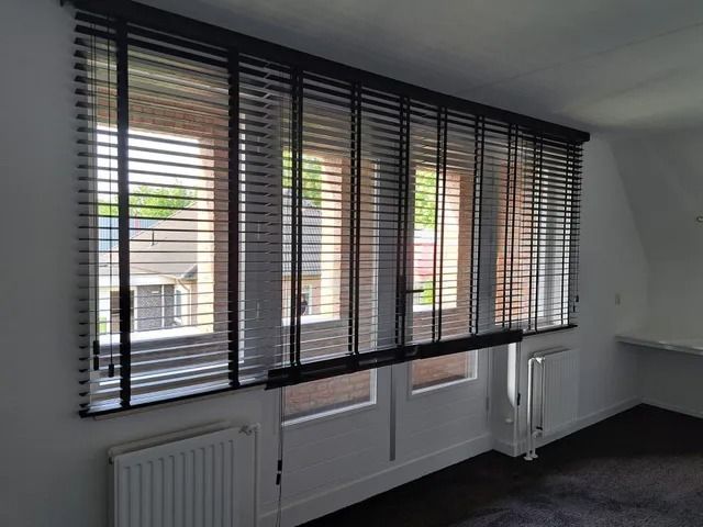 Lege kamer met grote ramen, zwarte jaloezieën en witte radiatoren onder de vensterbanken.