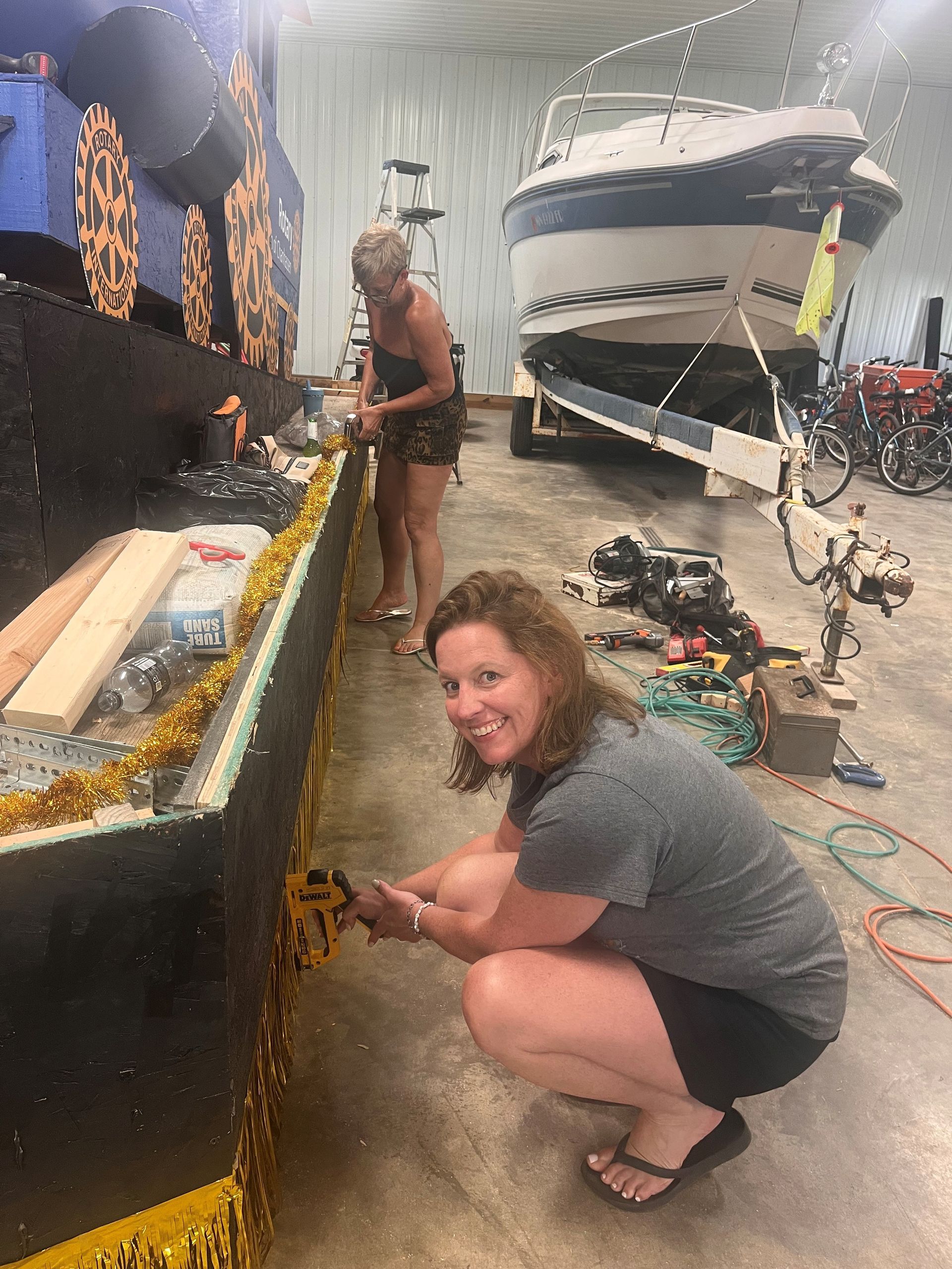 Woman crouching beside a decorated boat in a garage, smiling near a white-and-blue boat on a trailer.