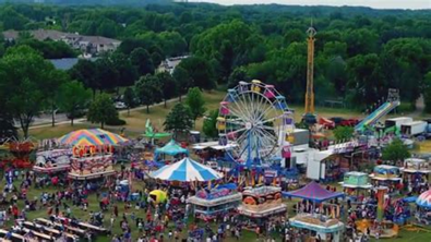 Aerial view of a crowded outdoor fair with a Ferris wheel, colorful tents, rides, and many visitors