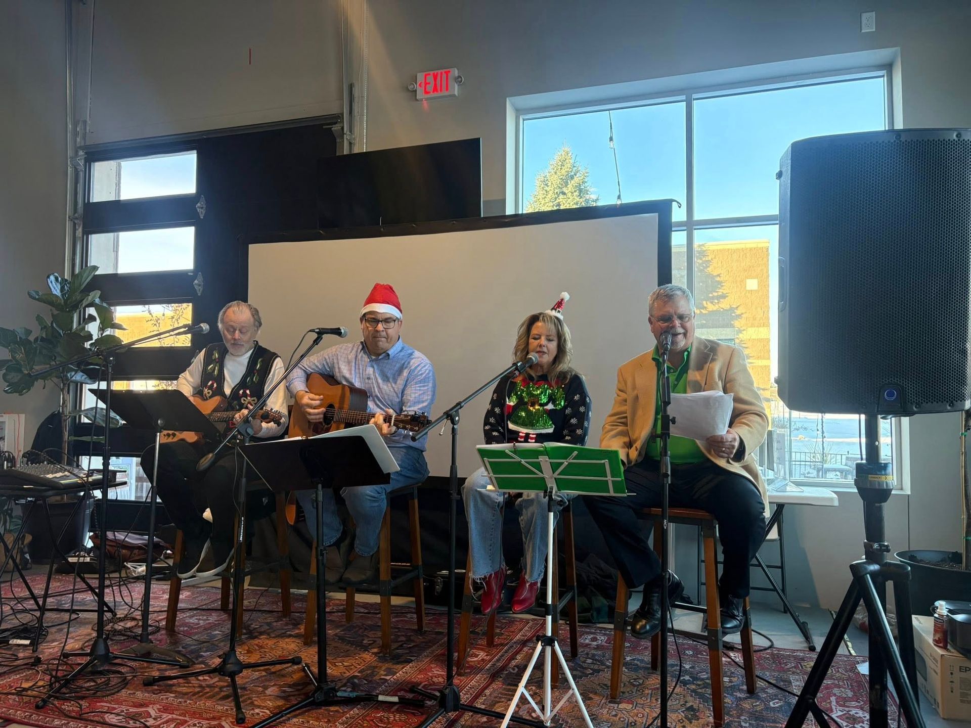 Four musicians performing indoors, with guitars, sheet music stands, and holiday hats near a large window