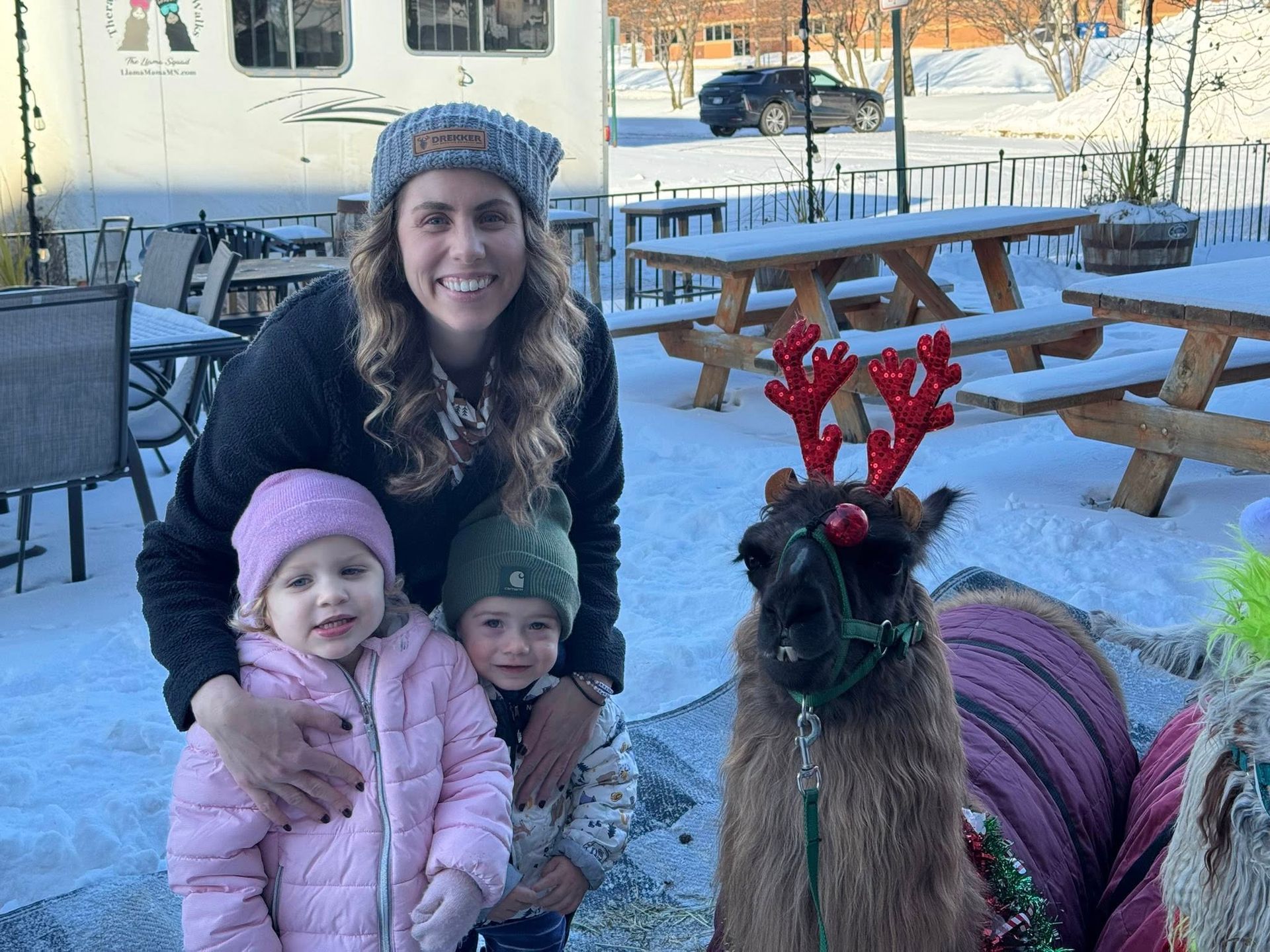 Two children with a smiling adult and a llama in reindeer antlers at a snowy outdoor café