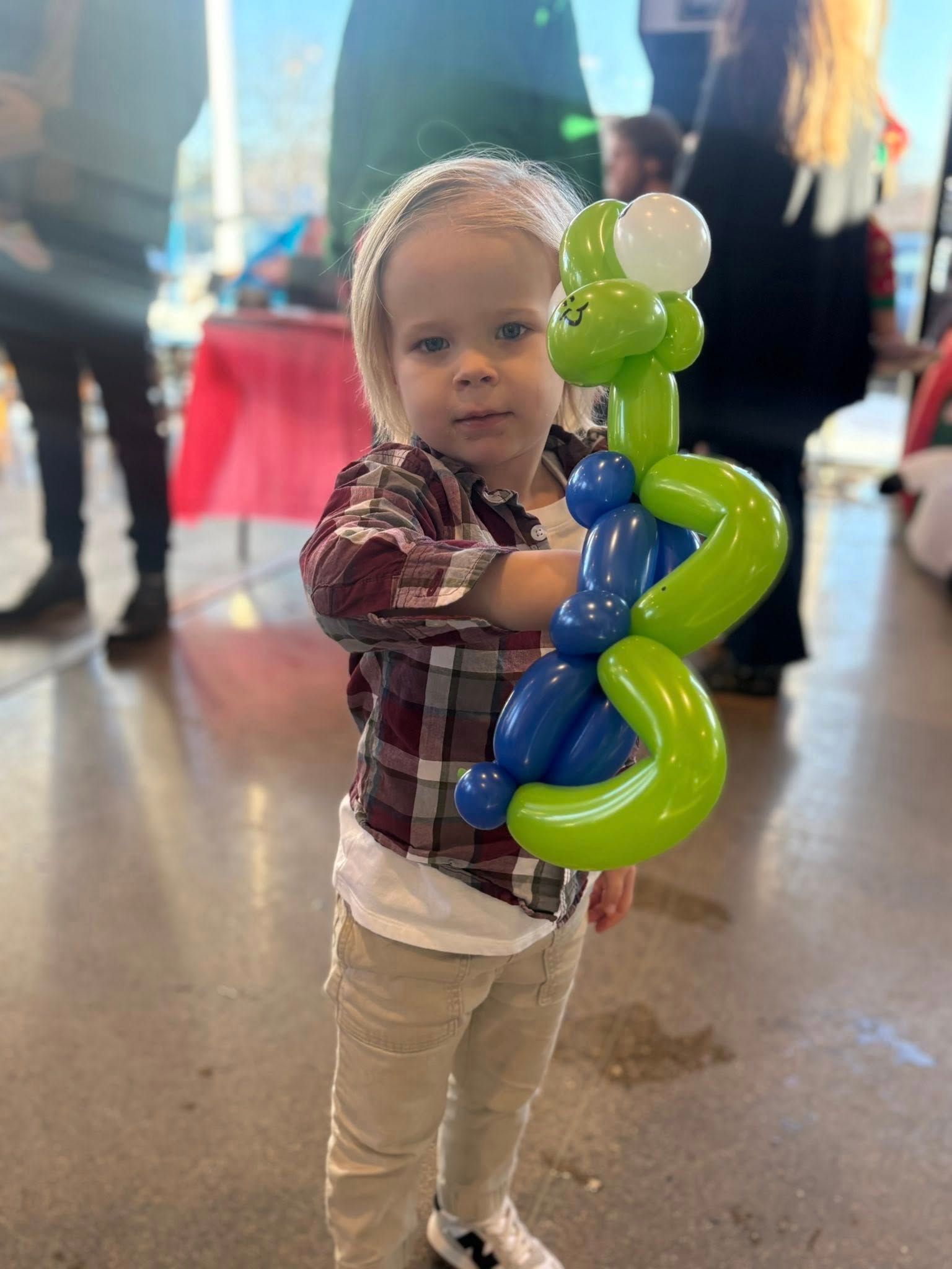Toddler holding a green and blue balloon sculpture indoors, looking at the camera.