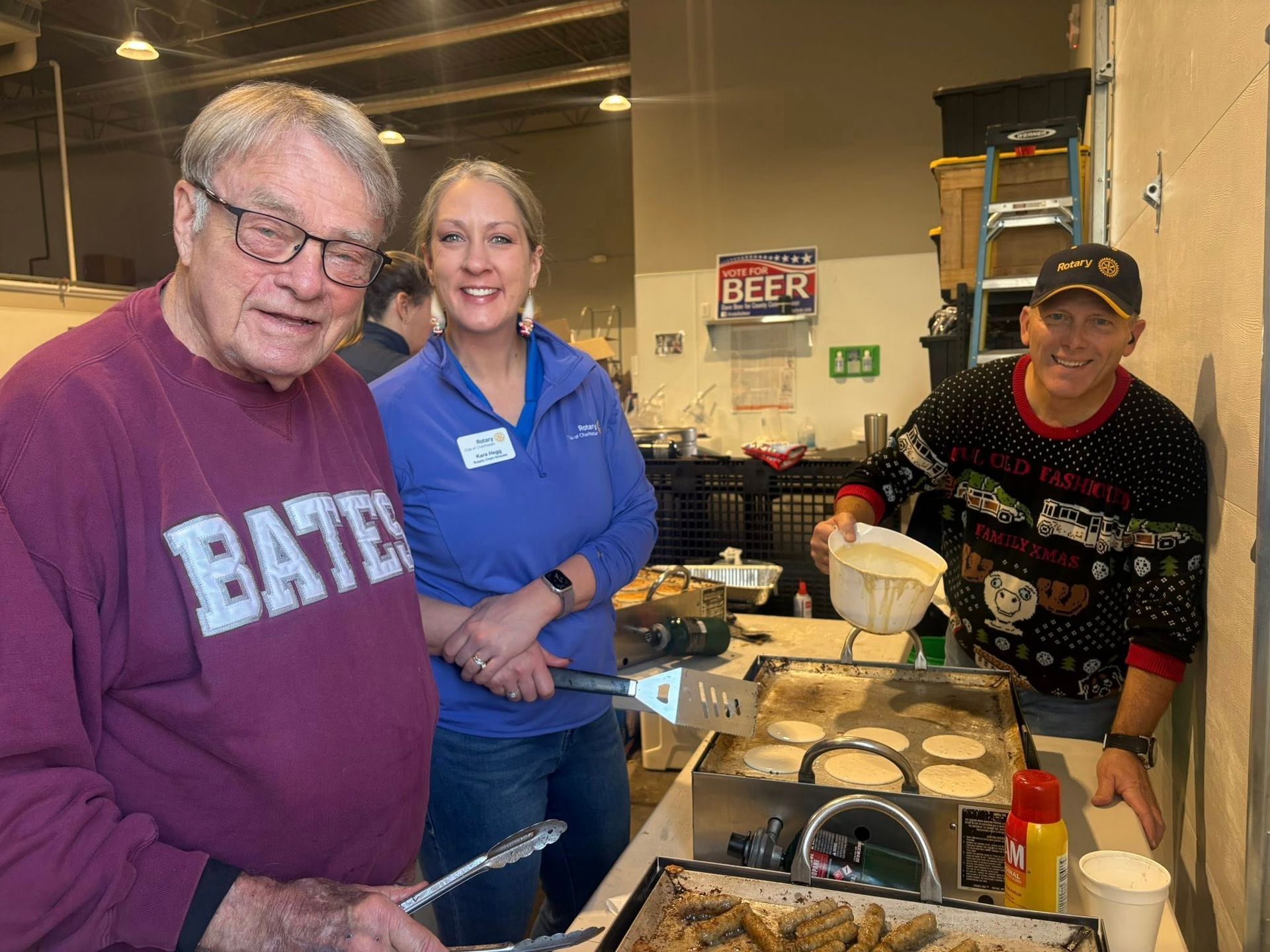 Three people in an industrial kitchen smile beside a worktable with food and dishes.