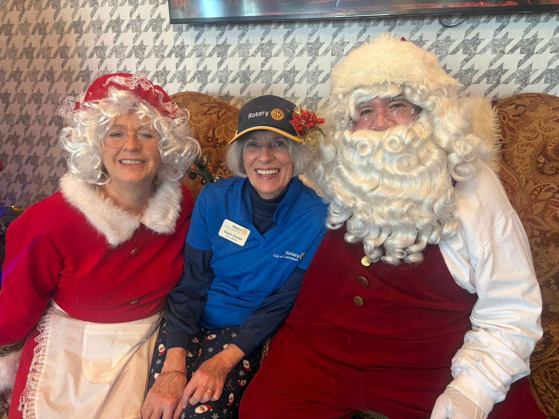 Three people in festive holiday costumes smiling together indoors, including Santa Claus.