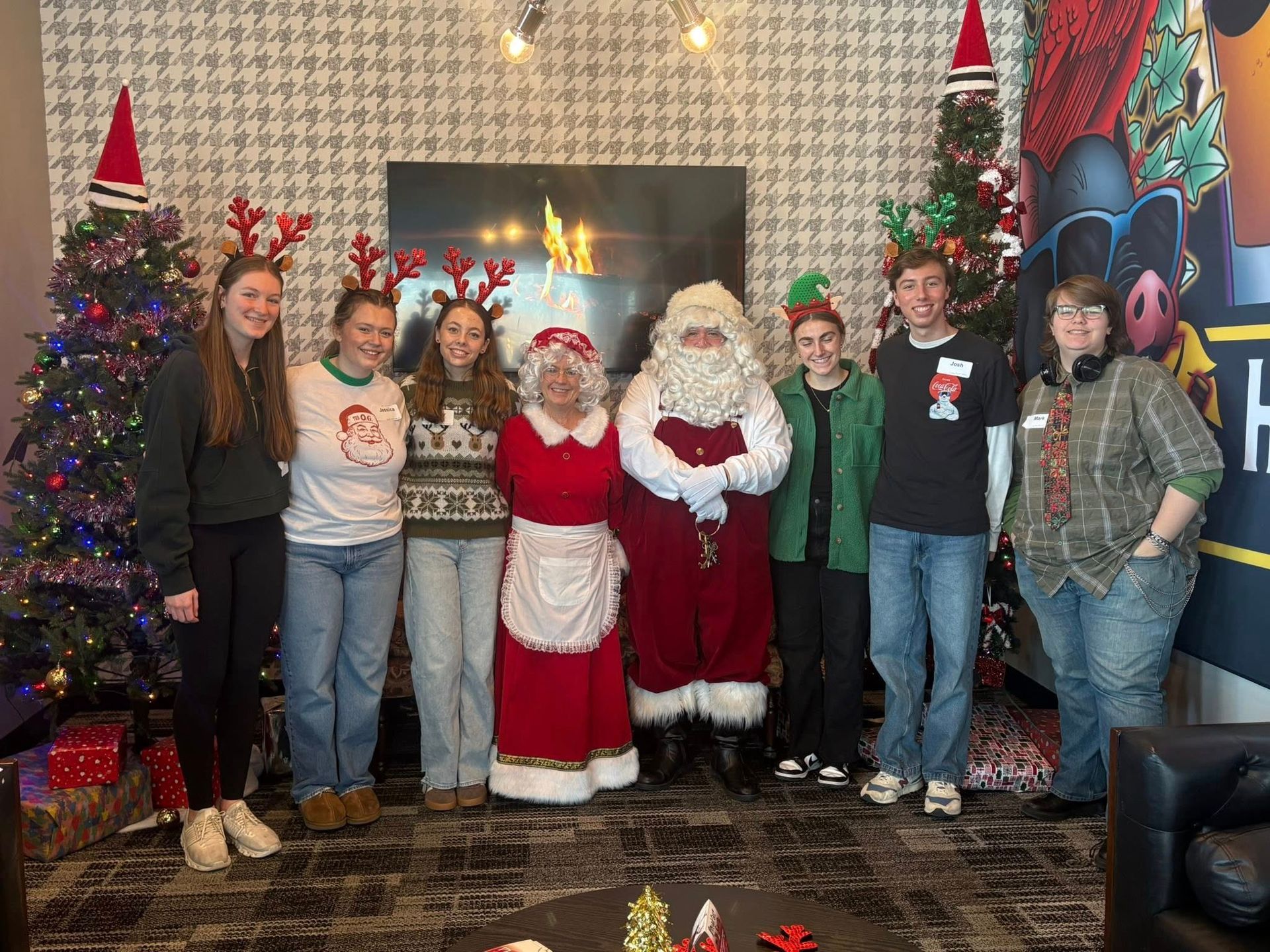 Group posing with Santa and Mrs. Claus in a decorated living room beside a fireplace and Christmas tree