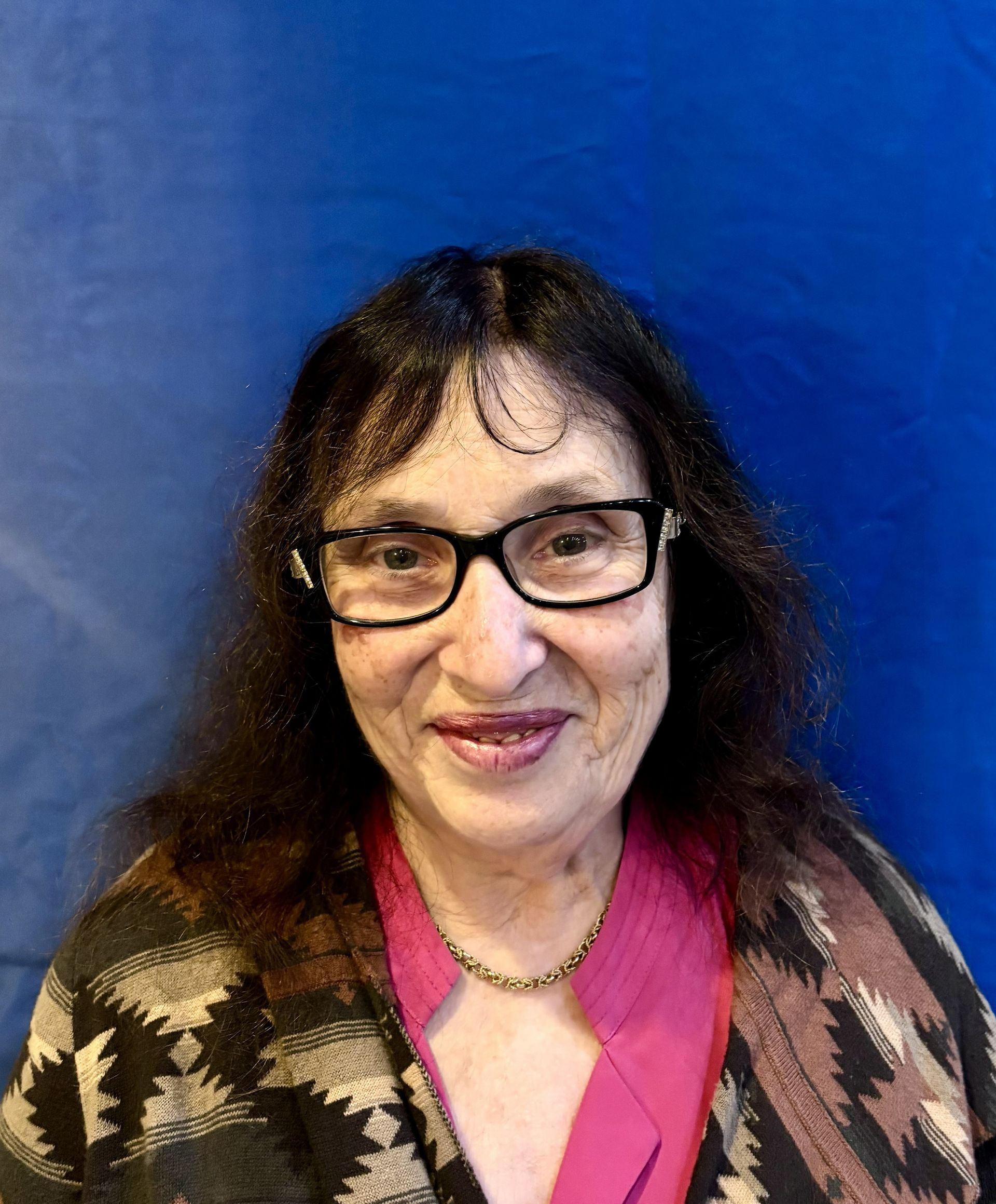 Smiling person with glasses and dark hair in a pink top, standing against a blue background