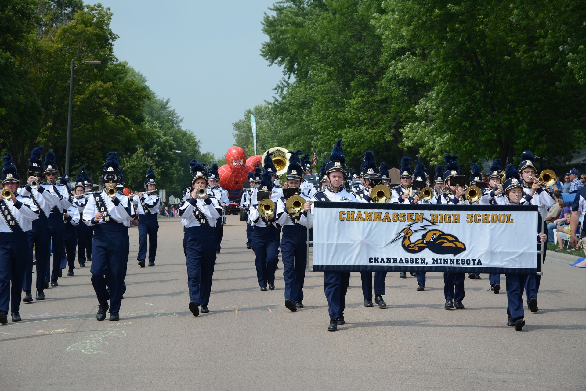 Marching band parade on a tree-lined street, with banners and uniforms leading the group.