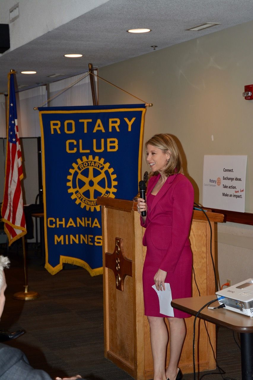 Woman speaking at a podium beside a Rotary Club banner in a meeting room