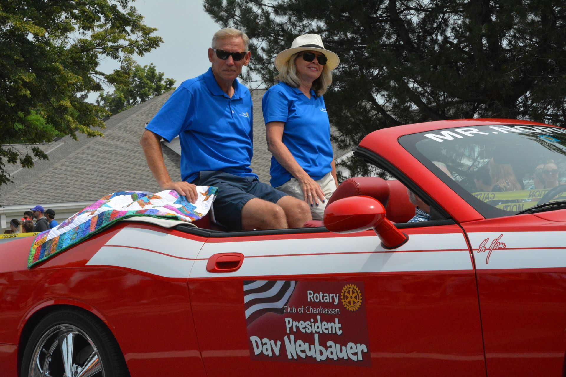 Two people in blue shirts sit in a red convertible decorated for a parade.