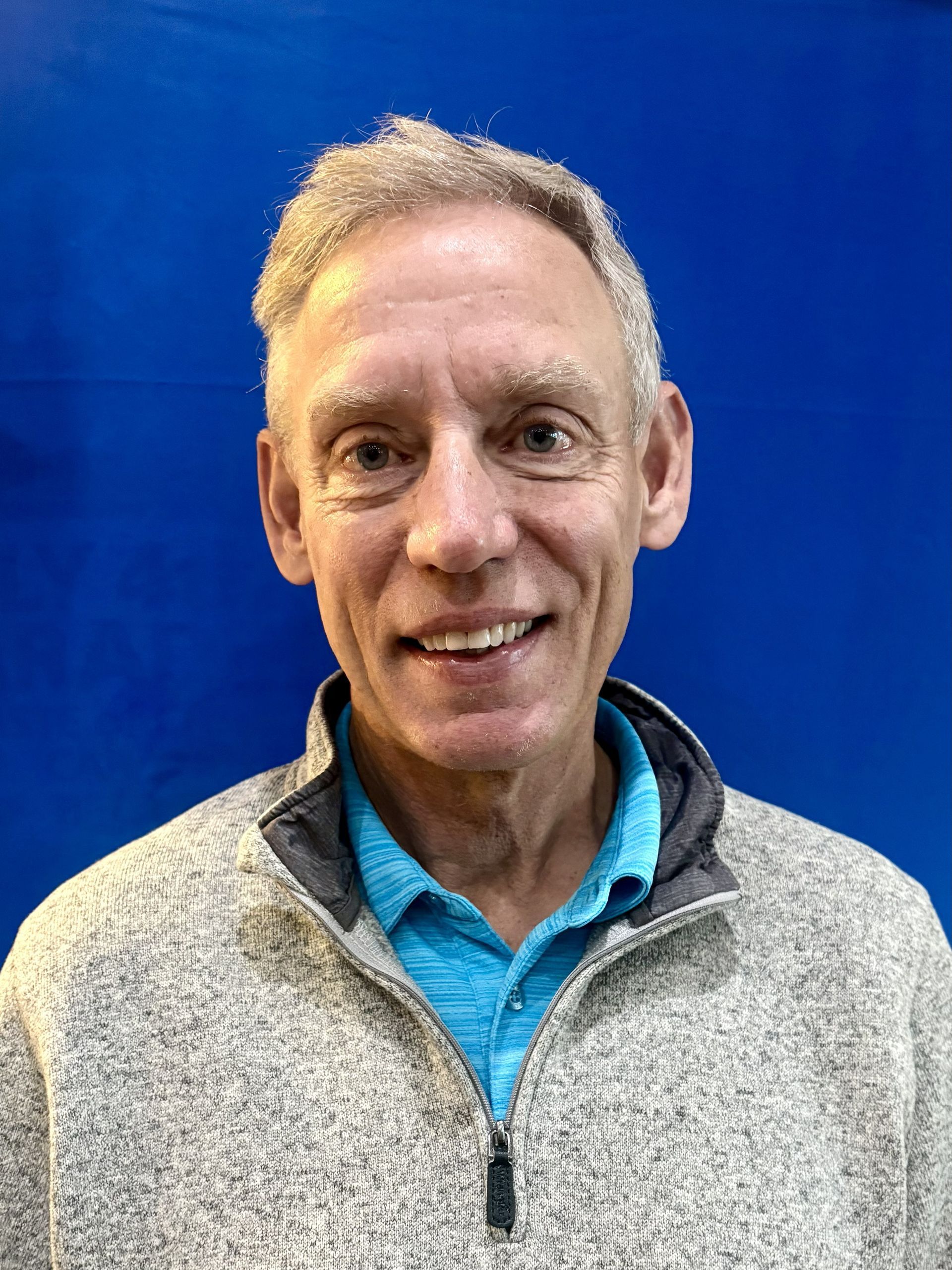 Smiling man in a gray sweater and blue shirt standing against a blue background