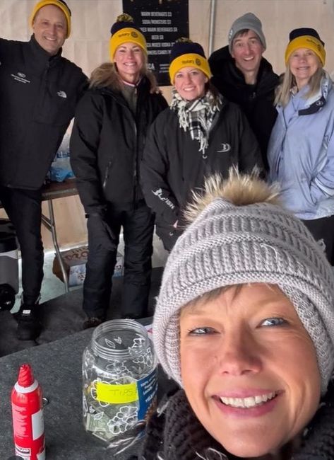 Five smiling people in winter hats posing indoors, with a selfie in the foreground