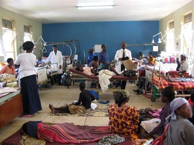Crowded hospital ward with patients on beds and people standing, blue walls and bright windows