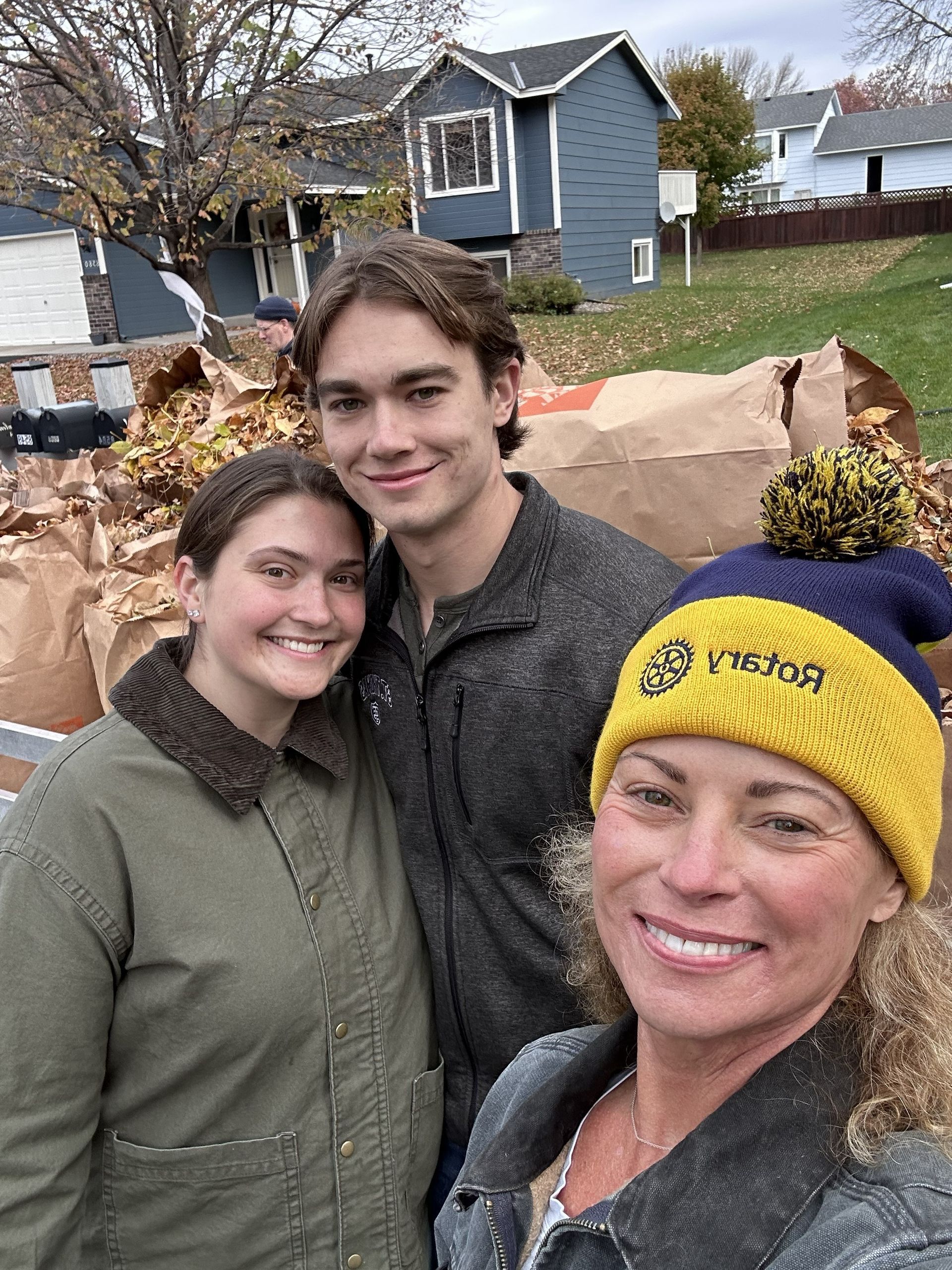 Three people smiling, posed near bags of fall leaves. One wears a yellow and blue hat. Houses in the background.