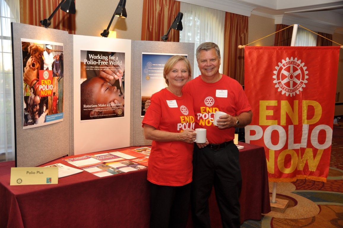 Two people in red shirts pose at a booth with anti-polio posters and a bright red “END POLIO NOW” banner.
