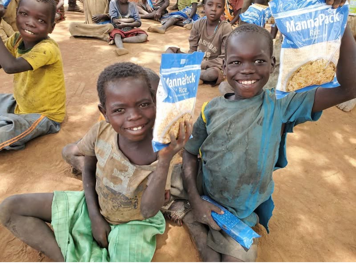 Children smiling and holding up food aid packets outdoors on dusty ground