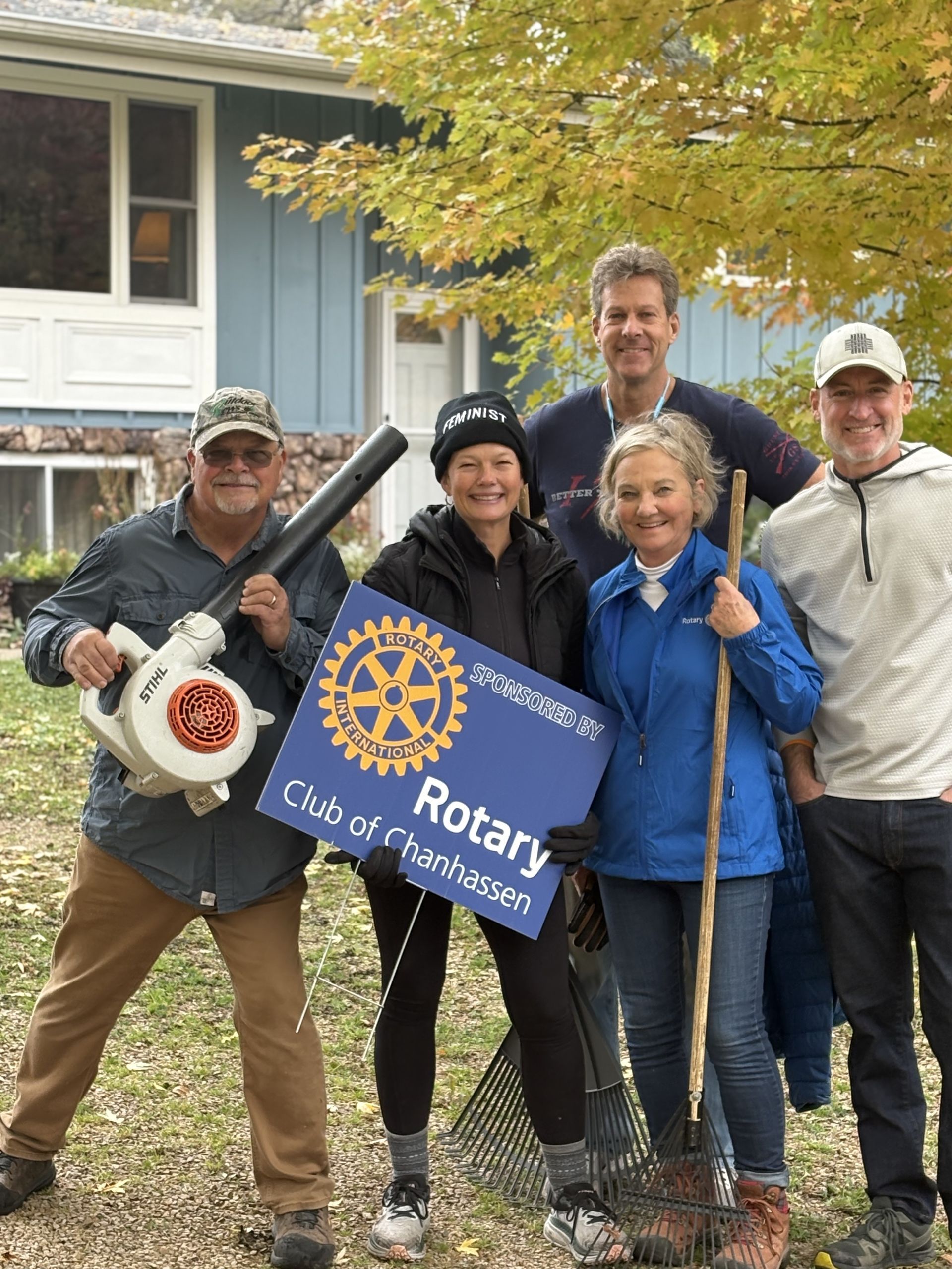 Five people smiling outdoors, holding a blue Rotary banner in front of a house and fallen leaves.