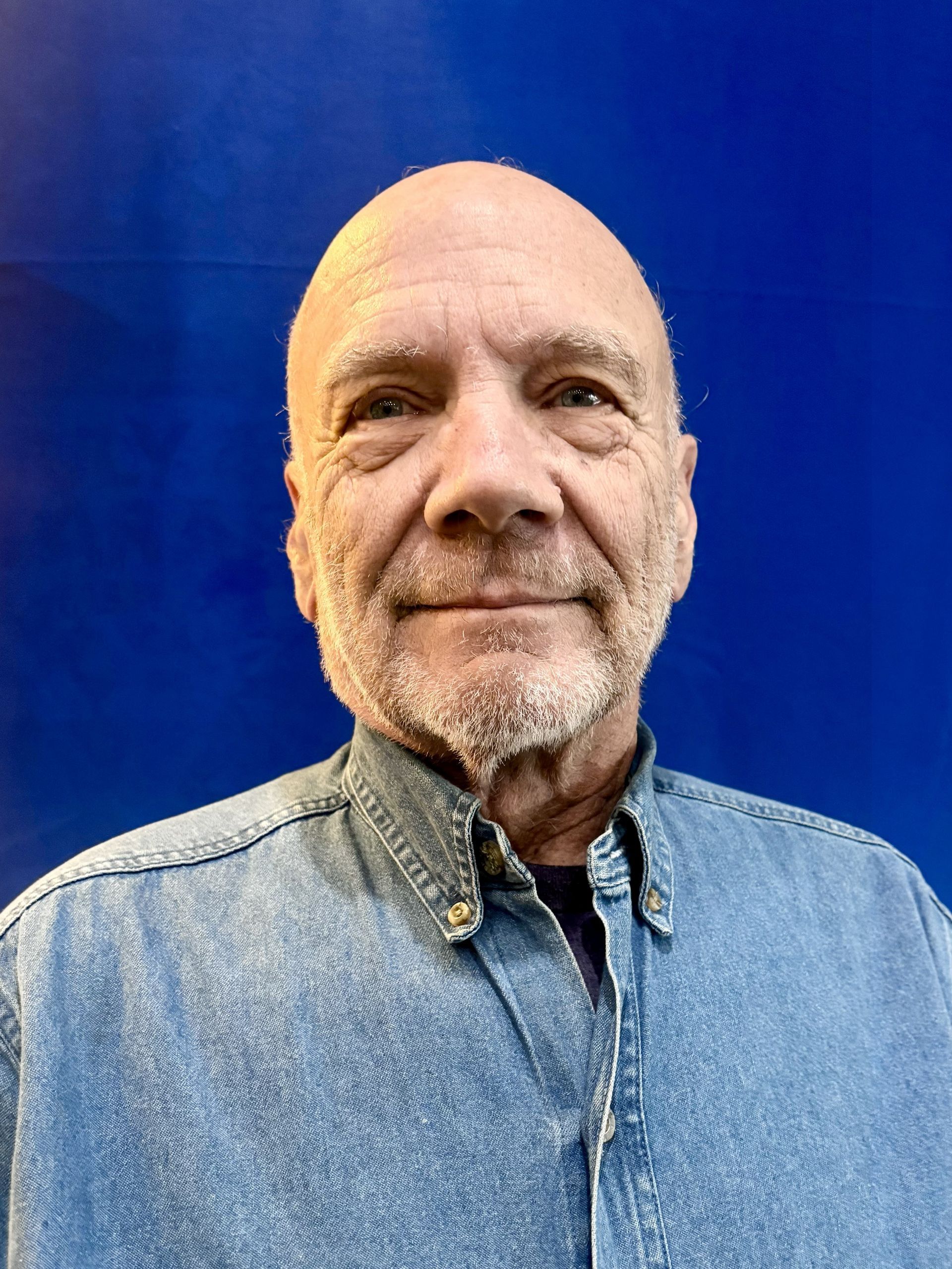 Portrait of a man in a denim shirt against a blue background, looking at the camera with a slight smile