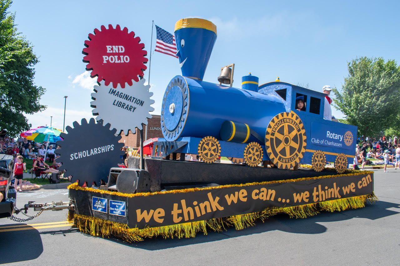 Blue train parade float with “we think we can” slogan and colorful balloons outdoors