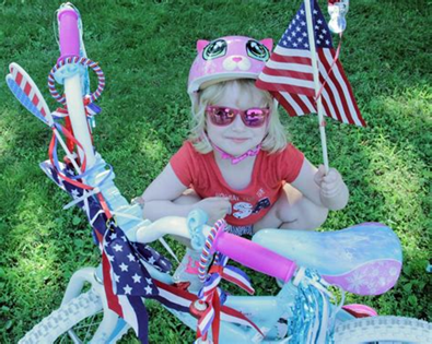 Child on a decorated bicycle holding an American flag, wearing a pink helmet and sunglasses on grass.