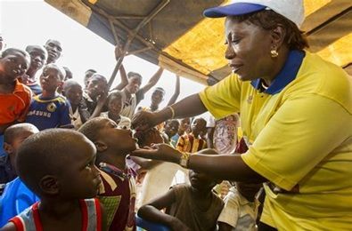 Woman administering oral vaccine to children in an outdoor setting. Many children watch.