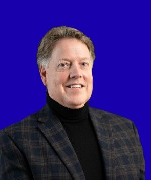 Studio portrait of a smiling person in a dark plaid blazer and black turtleneck against a blue background
