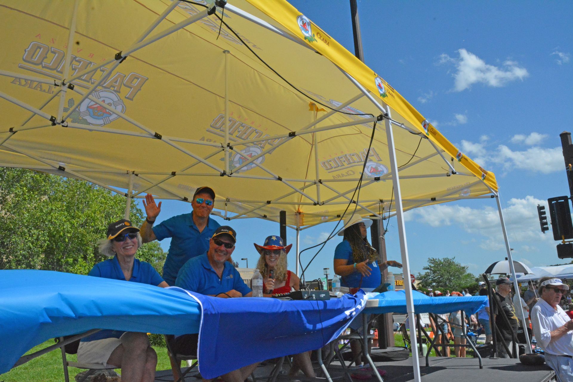 People under a yellow canopy at an outdoor event, with blue banners and a sunny sky.