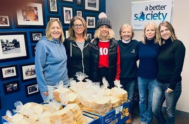Five women stand behind baskets of bread at a charity booth with a “better society” sign.
