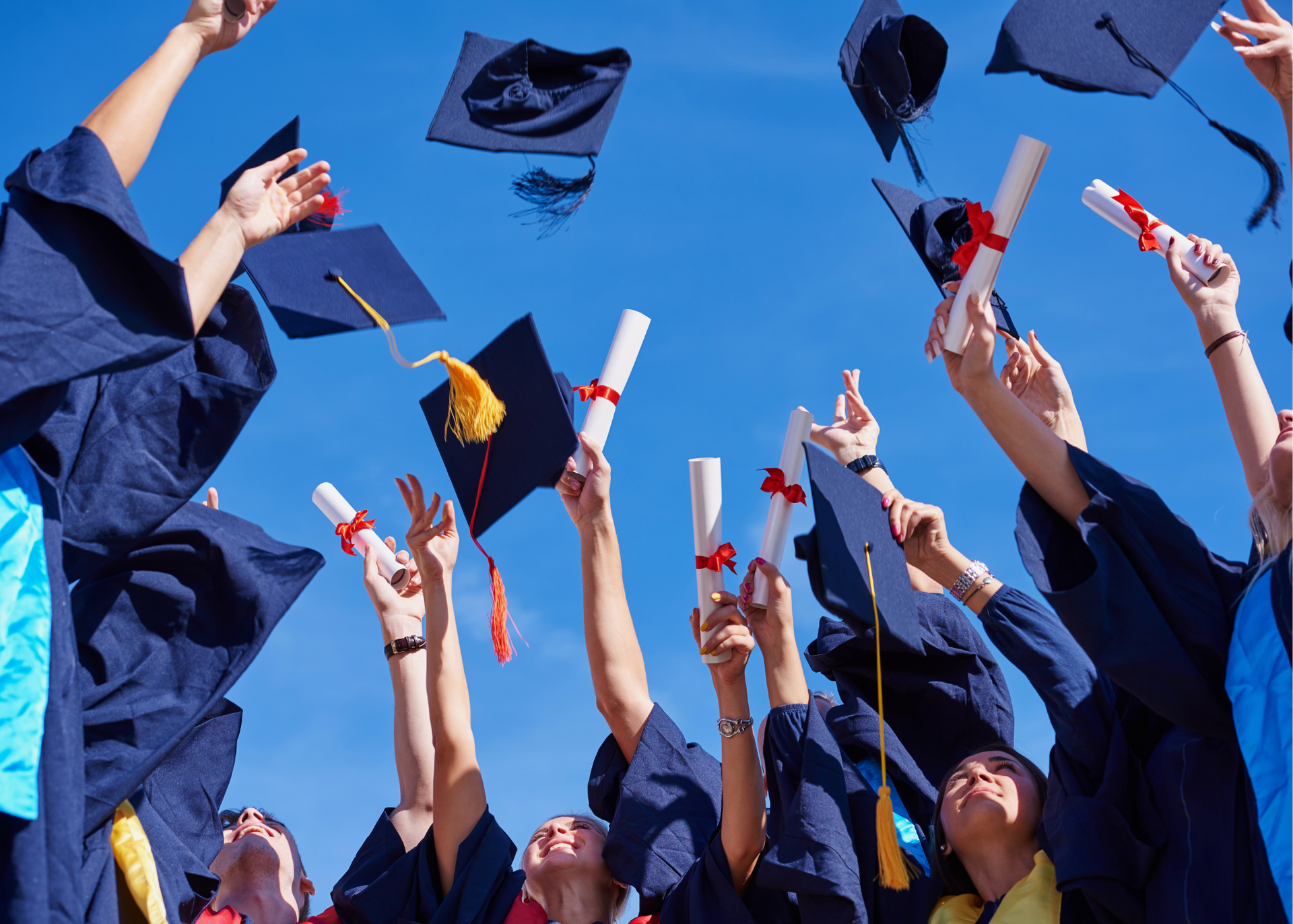 Graduates in blue gowns toss caps and diplomas into a bright blue sky