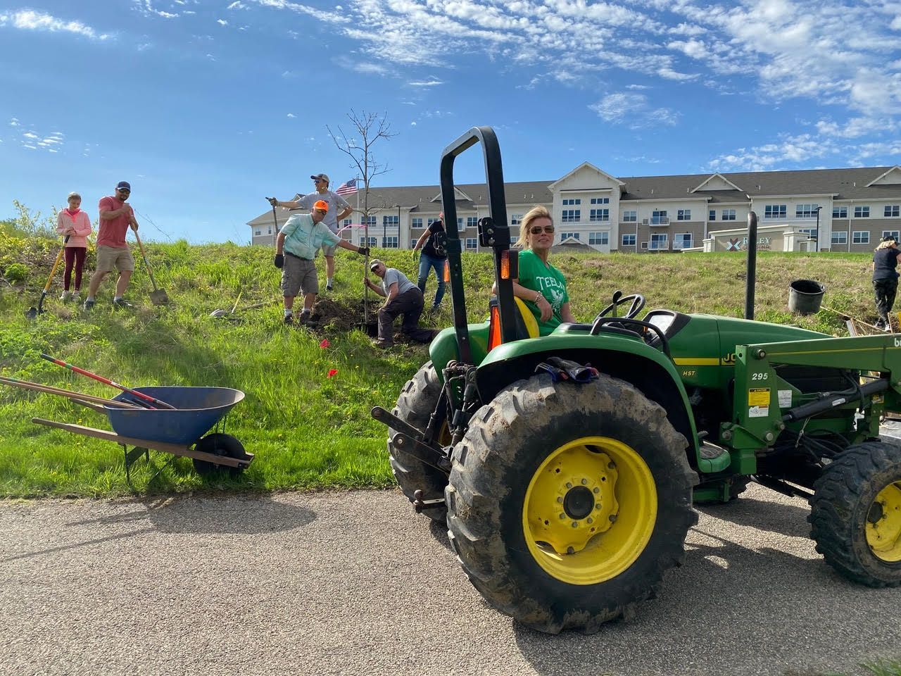 Green tractor with yellow wheels on a grassy slope, with people and a building in the background