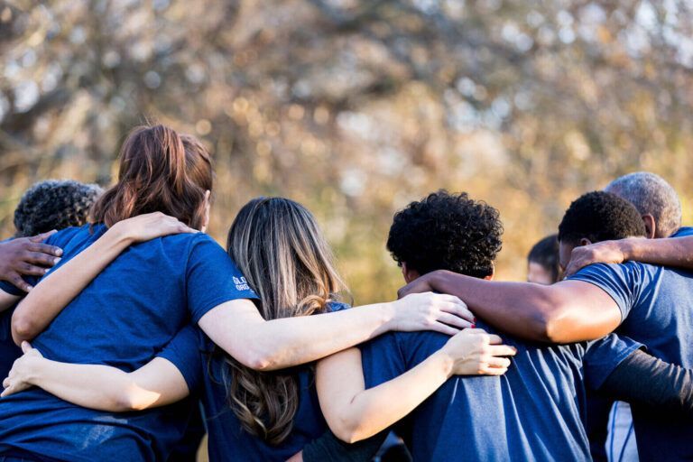 Group of people in blue shirts standing arm-in-arm outdoors with trees in the background