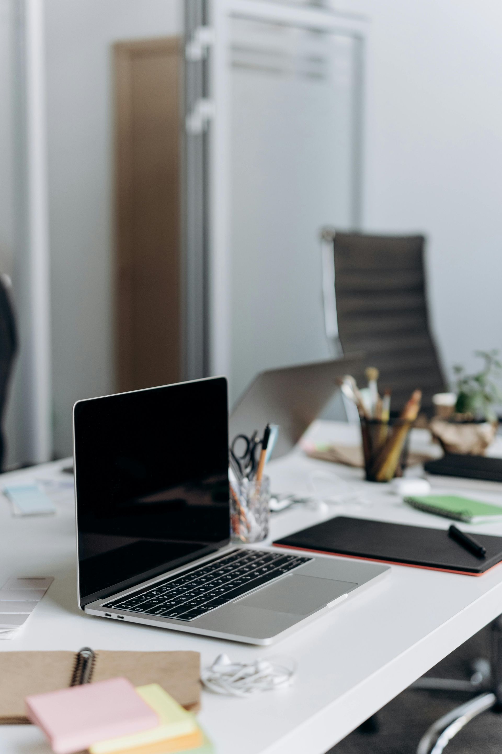 Laptop on a white office desk with notebooks, pens, and a chair in the background