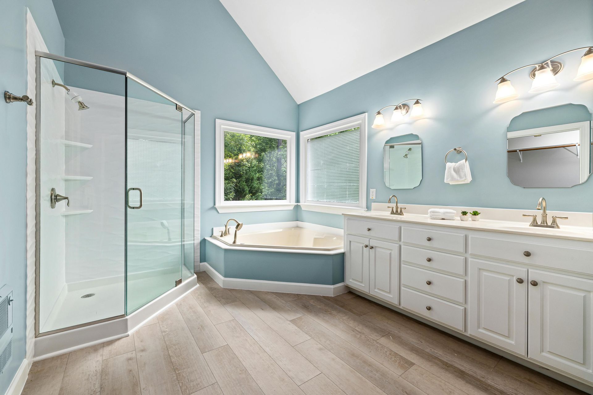Bright blue bathroom with glass shower, soaking tub, double vanity, and light wood floors.