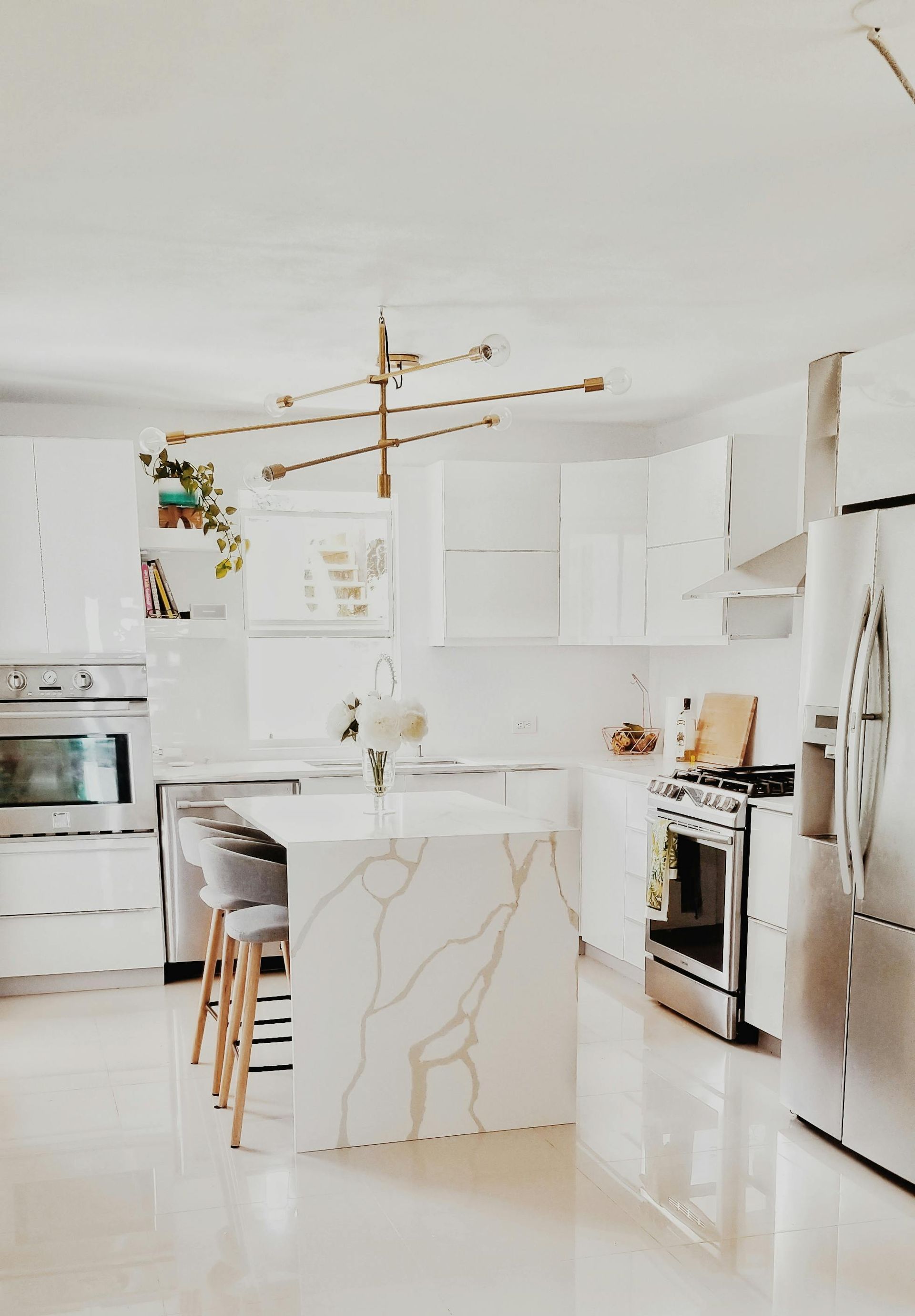 Bright white modern kitchen with marble island, gold pendant light, and stainless steel appliances.
