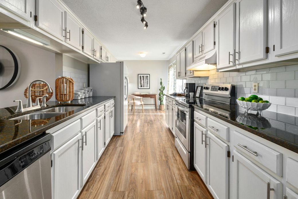 Bright galley kitchen with white cabinets, stainless appliances, wood floors, and a dining area beyond.