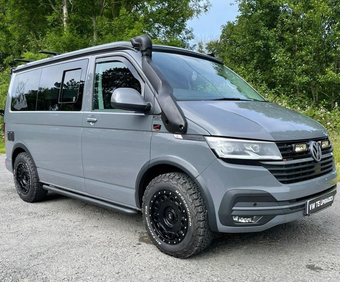 Grijze Volkswagenbus met zwarte offroadvelgen, snorkel en imperiaal staat geparkeerd op een grindweg.