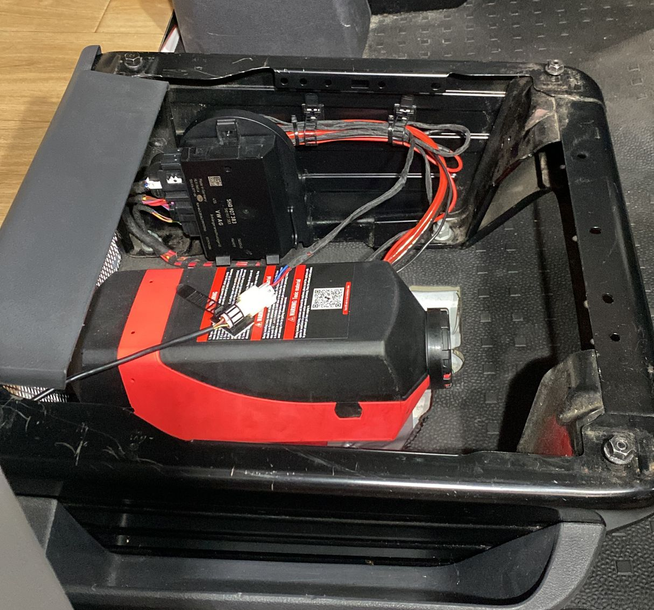 Dieselkachel geïnstalleerd onder een autostoel, met zichtbare zwarte en rode componenten en bedrading.