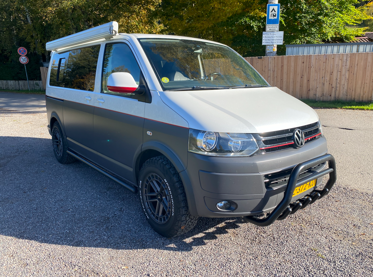 Witte en grijze camper met zwarte accenten en luifel geparkeerd op grind.