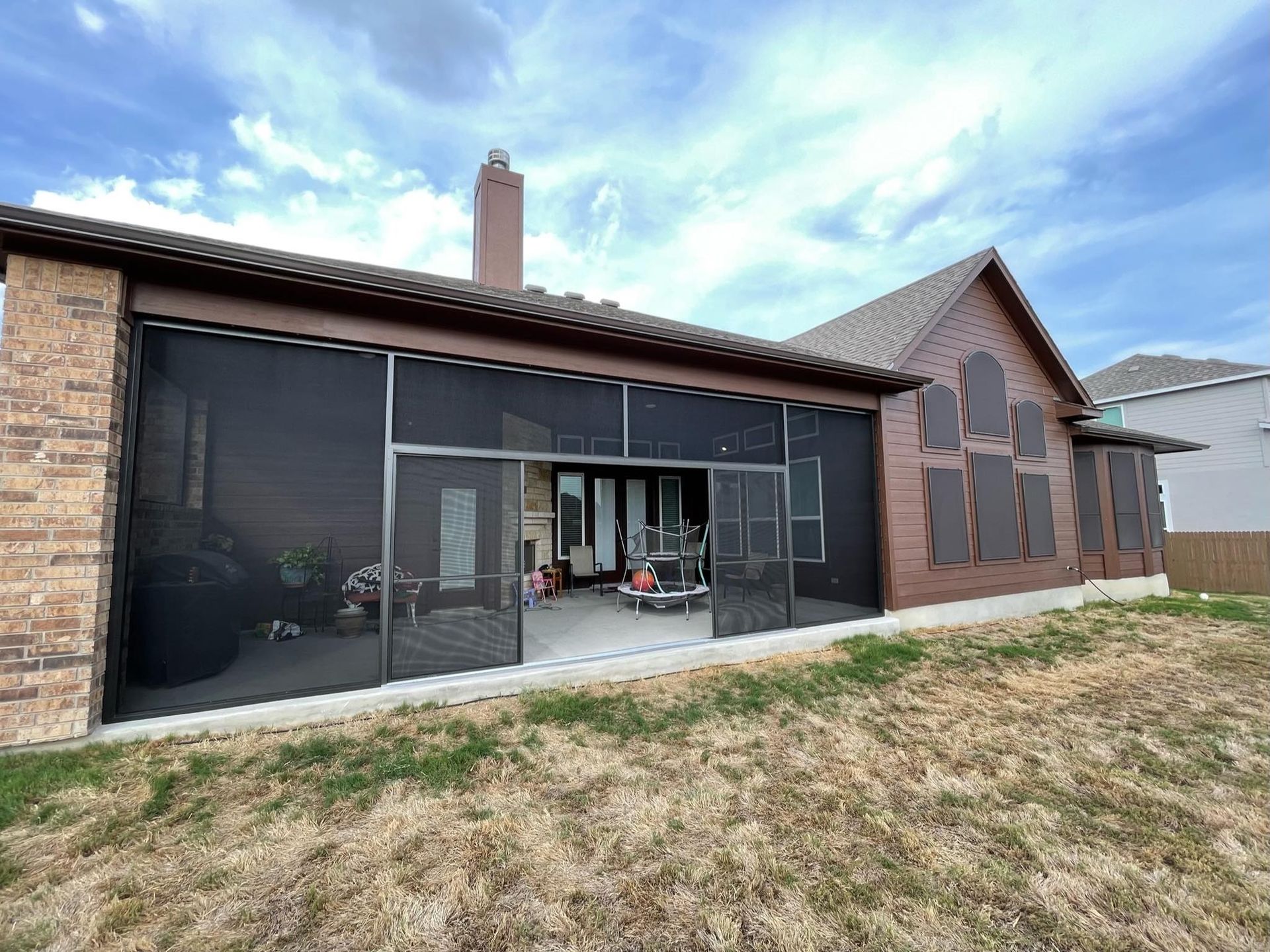 A brown brick house with a large screened-in porch and chimney, viewed from a dry grass lawn under a blue, cloudy sky.