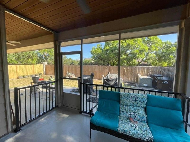 A screened-in patio with a teal sofa, a metal railing, and a view of a backyard with a wooden fence and trees.