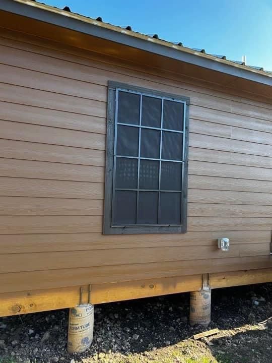 A rectangular window with a dark metal grid and screen set into the horizontal tan siding of a house on concrete pilings.