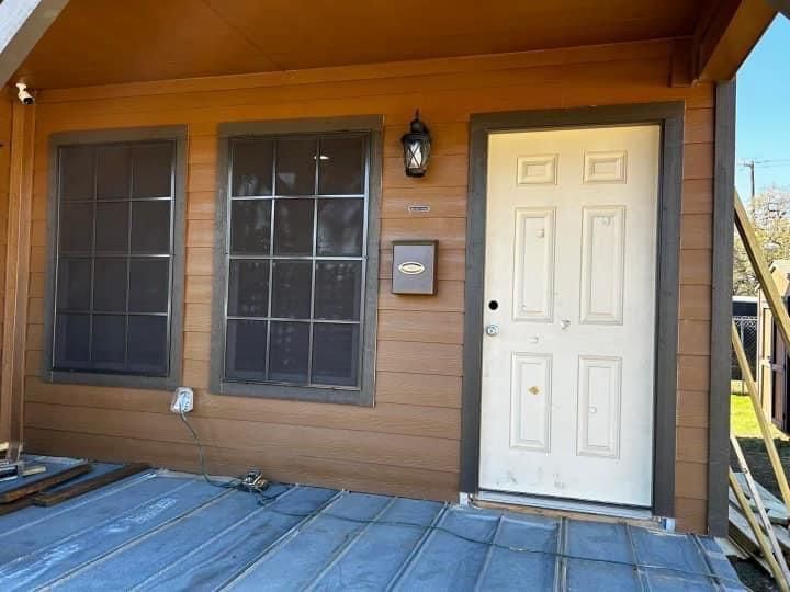 Exterior of a tan building with two dark-framed windows, a light-colored door, and a metal-paneled porch floor.