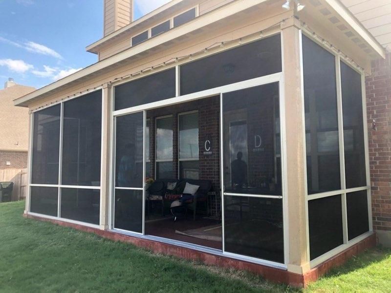 A beige screened-in porch with sliding doors on a brick house, overlooking a green lawn under a blue sky.