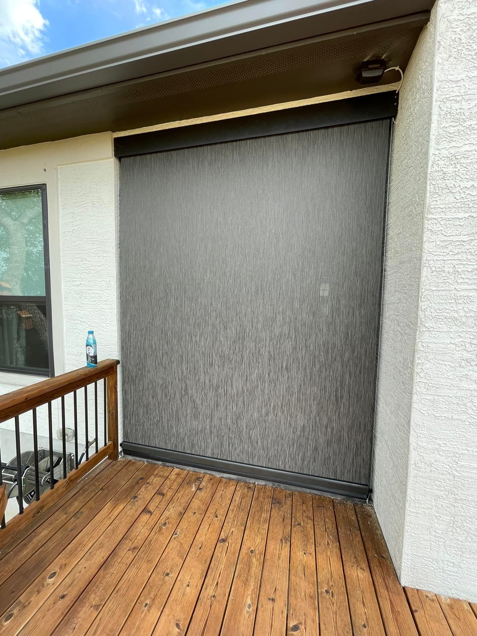 A dark gray outdoor roller shade is installed on a wooden deck under a porch roof next to a stucco wall.