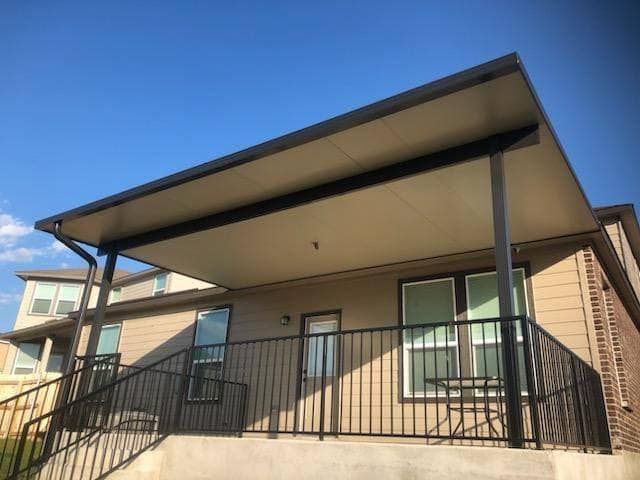 A patio with a dark metal-framed covered roof, black railing, and beige exterior siding against a blue sky.