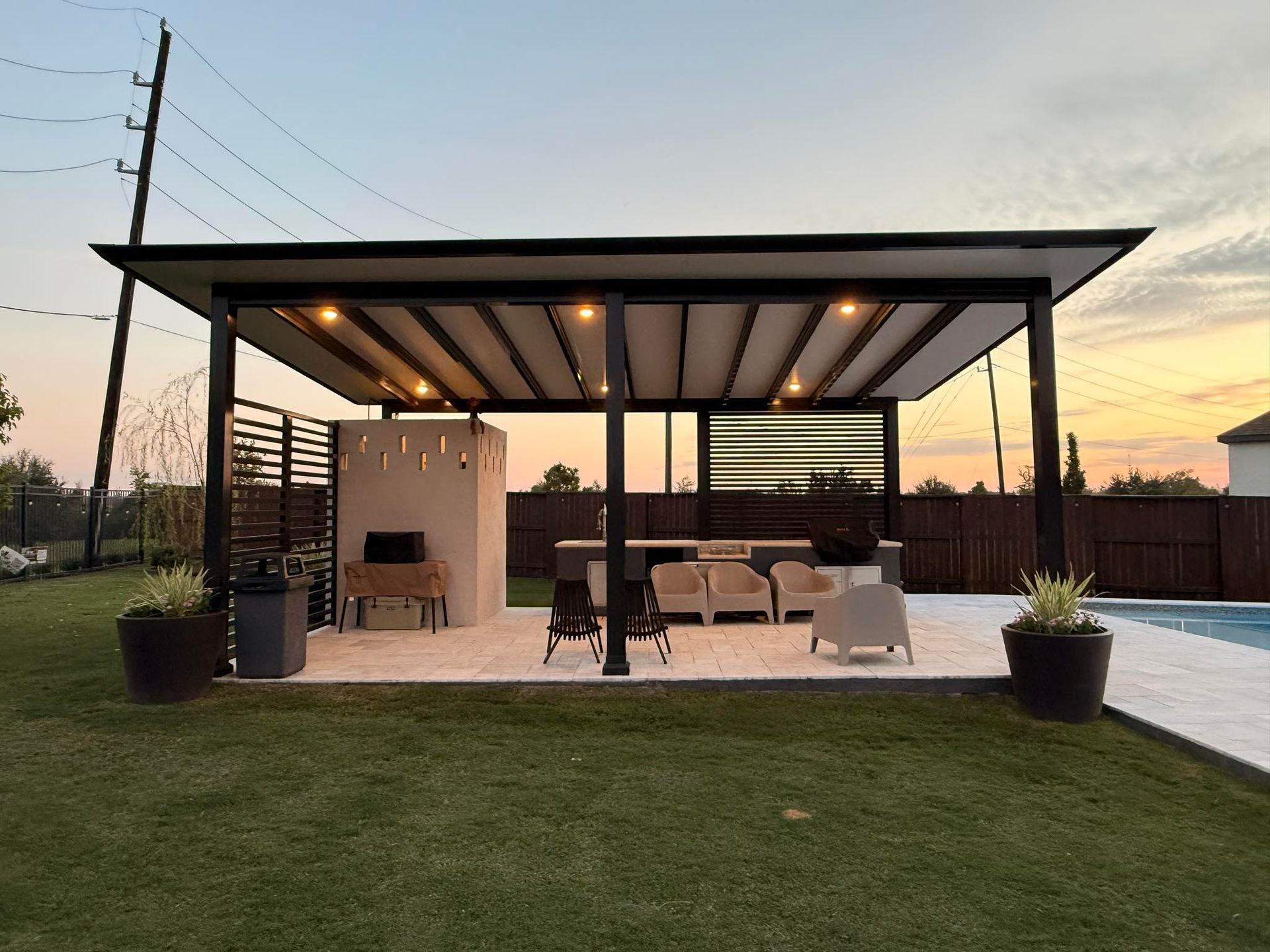 An outdoor covered patio with seating, a small kitchen area, and string lights, situated by a pool at sunset.