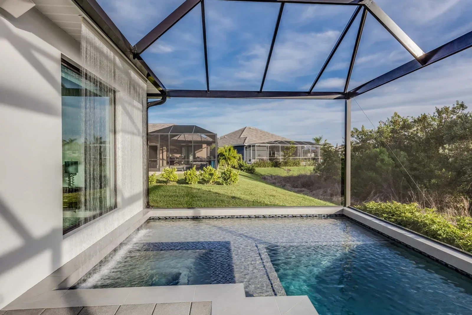 A small backyard swimming pool with an attached spa enclosed by a screened-in lanai, overlooking a grassy field and houses.